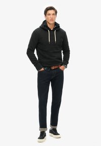 Sudadera negra con capucha y logo texturizado, camisa de cuadros azul debajo, jeans oscuros y zapatillas negras con suelas blancas.