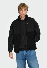 Giovane uomo con capelli biondi scompigliati, indossa un gilet di pile nero sopra una felpa nera e jeans blu, in piedi con una mano in tasca.