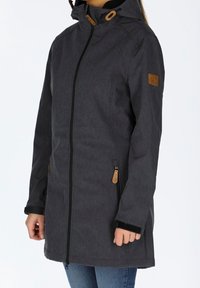 Personne portant une veste à capuche gris foncé avec des poches zippées et une bordure noire, associée à un jean bleu sur un fond uni.