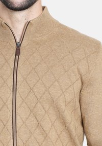 Sudadera cremosa con cremallera que presenta un patrón de rombos, cuello acanalado y cremallera marrón. La tela parece suave y texturizada.