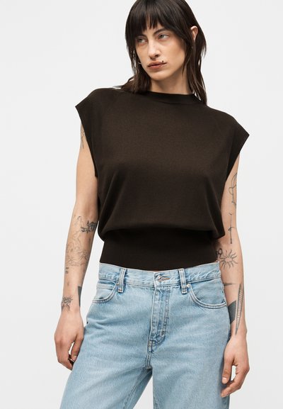 Personne aux cheveux foncés, tatouages sur les bras, portant un haut noir sans manches et un jean bleu clair taille haute sur un fond uni.