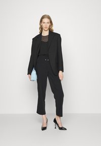 Blazer preto ajustado sobre uma blusa preta com bolinhas, combinado com calças largas pretas e sapatos de salto pretos. Segurando uma mala texturizada azul clara.