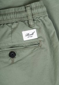 Close-up of green pants waistband with elastic, belt loop, back pocket with black button, and white stitched brand label "Reell est. 1997."