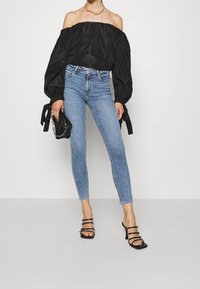 Top negro sin hombros con mangas voluminosas, combinado con unos pantalones vaqueros ajustados de color azul claro y sandalias de tiras negras. Los accesorios incluyen un pequeño bolso de mano negro.