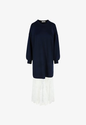 Námornícky svetrový šaty s dlhými rukávmi a okrúhlym výstrihom, preložené cez bielu krajkovú sukňu s kvetinovými vzormi a splývavým lemom.