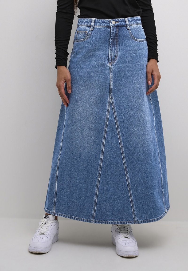 Falda maxi de mezclilla en azul claro con cintura ajustada, forma en A, bolsillos frontales y costuras de contraste. Combinada con zapatillas blancas.