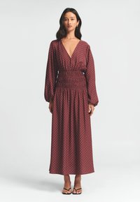 Hnědá maxi šaty s dlouhými rukávy, bílými puntíky, hlubokým výstřihem do V, smockovaným pasem a volnou sukní; nošeny s černými sandály.