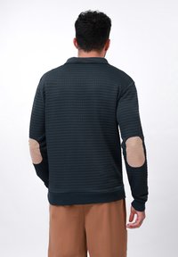 Mørkeblå sweatshirt i waffle-strikk med beige albueputer, rund halsudskæring og ribbede manchetter. Relaxed fit, parret med lysebrune shorts.
