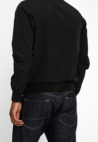 Veste bomber noire avec une texture lisse, des poignets côtelés et un ourlet. Associée à un jean en denim foncé avec des coutures jaunes et des poches arrière.
