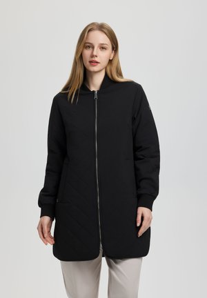 Junge Frau mit glattem blondem Haar trägt einen langen schwarzen Reißverschlussmantel mit gerippten Bündchen und beige Hose, steht vor einem grauen Hintergrund.