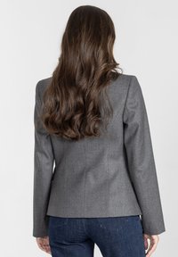 Kobieta z długimi falującymi brązowymi włosami nosi dopasowaną szaro-skalną marynarkę i ciemnoniebieskie dżinsy, sfotografowana od tyłu na jednolitym białym tle.