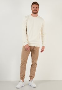 Crèmepullover met ronde hals, gecombineerd met lichtbruine tapered pants. Witte sportschoenen maken de outfit compleet.