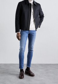 Černá zipová bunda s límcem; krémový svetr pod ní; modré úzké džíny; lesklé tmavě hnědé mokasíny; minimalistický design a textury.