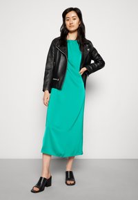 Vestido midi teal em tecido suave, em camadas sob uma jaqueta de motociclista de couro preta. Mules de salto bloco aberto pretas completam o visual.