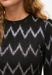 Mujer con un top negro texturizado con un patrón de zigzag plateado brillante, con cabello rubio rizado parcialmente visible, fondo neutro.