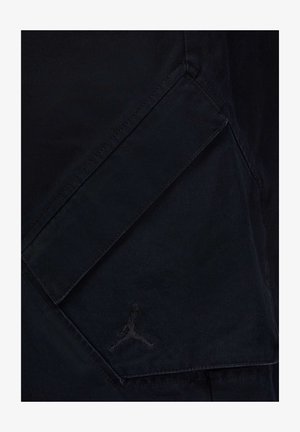 Czarna tkanina o matowej fakturze, z ukośną kieszenią i wyszywaną w ciemnym kolorze logo Jordana, podkreślająca minimalistyczny design.