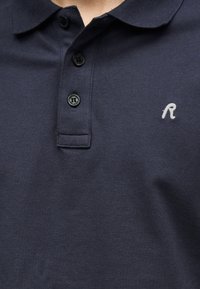 Μπλε ναυτικό πόλο πουκάμισο με τρία μαύρα κουμπιά και μικρό λευκό κεντημένο γράμμα "R" στο αριστερό στήθος.