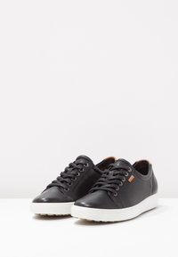 Baskets en cuir noir avec semelles en caoutchouc blanches, bout rond, six œillets et détails subtils de la marque en beige sur le côté.