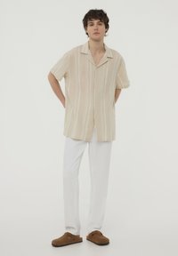 Camicia beige a maniche corte con bottoni, con strisce verticali, abbinata a pantaloni bianchi larghi e scarpe marroni senza lacci.
