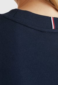 Námorně modrý svetr z látky s vroubkovaným výstřihem. Malý červený a bílý pruh zdobí límec, což naznačuje detail značky.