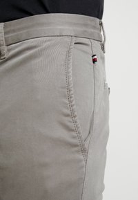 Närbild av ljusgrå byxor med midjeband och sidoficka som har en liten tygetikett i röd, vit och blå.