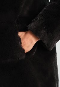 Svart fuskpälsjacka med en mjuk textur, med en sidficka och breda ärmkanter. Tyget verkar tätt och pluffsigt.