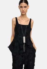 Kobieta z włosami zaczesanymi do tyłu, ubrana w czarny, teksturowany kombinezon bez rękawów oraz długi srebrny naszyjnik z wisiorkiem, z rękami w kieszeniach, na neutralnym tle.