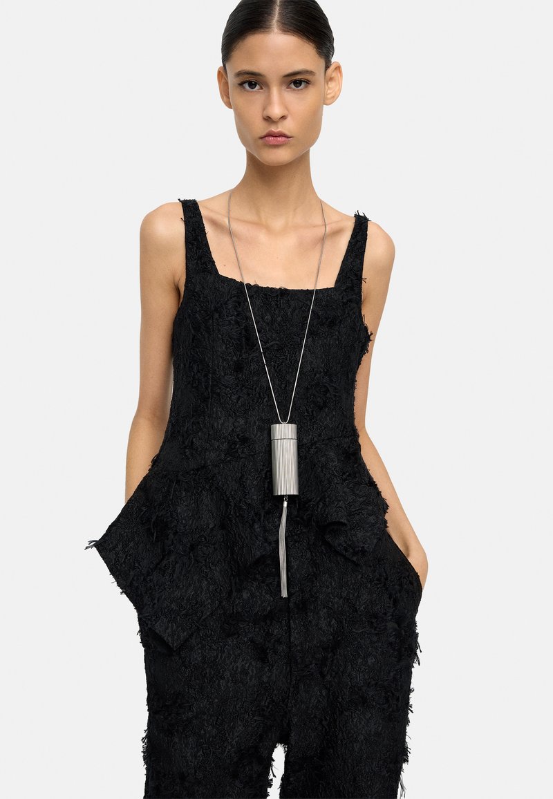 Kobieta z włosami zaczesanymi do tyłu, ubrana w czarny, teksturowany kombinezon bez rękawów oraz długi srebrny naszyjnik z wisiorkiem, z rękami w kieszeniach, na neutralnym tle.