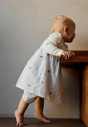 Un tout-petit se tenant sur la pointe des pieds, s'accrochant à une table en bois, portant une robe blanche avec de petites cerises rouges et un pantalon assorti.