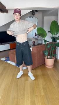 Lys beige t-skjorte, svarte denimshorts og hvite joggesko. Innendørs setting med tregulv og grønne planter i bakgrunnen.