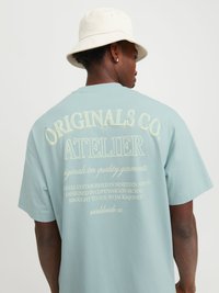 T-shirt en coton bleu clair avec un grand imprimé textuel au dos, avec des accents crème. Manches courtes et coupe décontractée. Chapeau seau au-dessus.