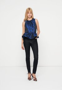 Μπλε σατέν τοπ με πεπλούμ και στρογγυλή λαιμόκοψη, σε συνδυασμό με μαύρο στενό τζιν και μαύρα πέδιλα με λουράκια, σε λευκό φόντο.