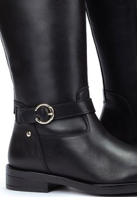Botas de cuero negro hasta la rodilla con una textura suave, que cuentan con una correa de hebilla circular plateada y un tacón bajo y apilado.