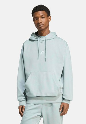 Junger Mann steht, trägt hellblauen Kapuzenpullover und passende Hose, Hände entspannt seitlich, neutraler Hintergrund.