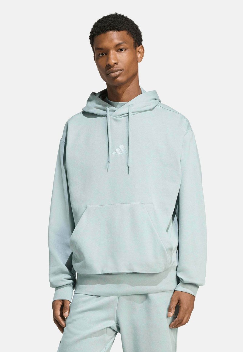 Jeune homme debout portant un sweat à capuche bleu clair et un pantalon assorti, les mains détendues le long du corps, fond neutre.