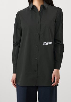 Vrouw draagt een lange zwarte blouse met knopen en het logo "KARL LAGERFELD JEANS" op de linkerborstzak, gecombineerd met donkerblauwe broek.