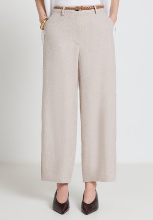 Pantaloni beige a gamba larga con tasche frontali, abbinati a una cintura marrone intrecciata e tacchi neri a punta, indossati con un top bianco e braccialetti dorati.