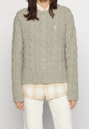 Personne portant un pull gris à torsades par-dessus une chemise à carreaux beige, associée à un pantalon blanc cassé, visible de la poitrine aux cuisses.