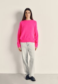 Sieviete stāv pie bēša sienas, valkājot gaiši rozā džemperi, gaiši pelēkas džinsas un melnas loafer kurpes, skatoties uz priekšu ar atslābinātām rokām.