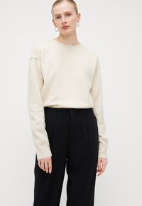 Pullover in maglia beige chiaro con collo rotondo e maniche lunghe, abbinato a pantaloni neri su misura con chiusura a bottone e pince.