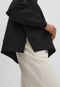 Felpa nera con orlo asimmetrico, caratterizzata da una consistenza morbida e polsini a costine, abbinata a pantaloni beige chiaro.