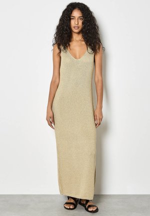 Kobieta o kręconych włosach, ubrana w beżową, dzianinową, długą sukienkę bez rękawów oraz czarne sandały, stojąca na tle białej ściany.