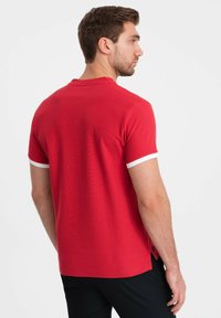Červená polo košile s krátkým rukávem a bílými akcenty, texturovaná tkanina, žebrovaný výstřih a boční větrací otvor pro prodyšnost, s volným střihem.