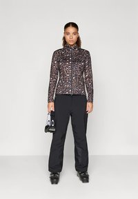 Casaco com fecho de correr com padrão de leopardo em tons de castanho e preto, combinado com calças de esqui pretas e botas de esqui pretas. Segura uma mala de mão com design de olhos.