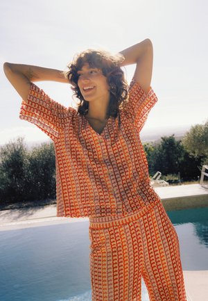 Femeie cu păr creţ, zâmbitoare, purtând un compleu portocaliu cu model, stând lângă o piscină, cu copaci în fundal, într-o zi însorită.