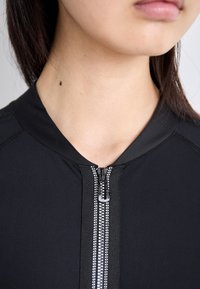 Schwarze angebotene Jacke mit glattem Stoff, Stehkragen, sichtbaren Nähten und einem silbernen Reißverschluss mit dekorativen Akzenten.