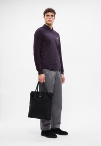 Schwarze Tote-Tasche mit zwei Griffen, aus Textilmaterial, kompaktes Design. Das Model trägt einen dunkelvioletten strukturierten Pullover und graue Hosen.