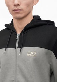 Άνδρας που φοράει μια δίχρωμη μαύρη και γκρι ζακέτα με φερμουάρ, κουκούλα και το λογότυπο EA7 Emporio Armani στο στήθος.