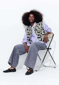 Gilet imprimé léopard par-dessus une chemise lilas à boutons, associé à un jean gris à jambes larges et des chaussures noires à clous. Chaise métallique.