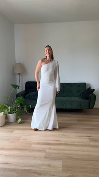 Vestido branco de um ombro, com corpete justo e saia fluida. Manga longa transparente com botões decorativos. Ao fundo, um sofá verde.